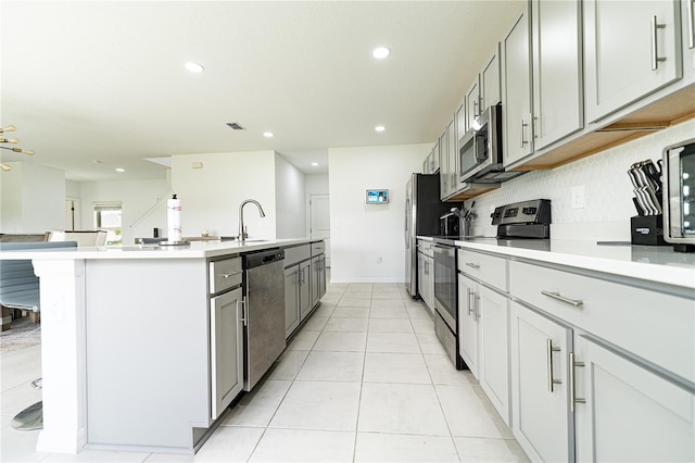 kitchen with stainless steel appliances, an island with sink, gray cabinets, backsplash, and light tile floors