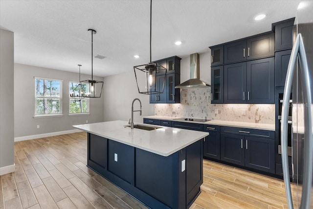 kitchen with black electric stovetop, sink, wall chimney range hood, a center island with sink, and decorative light fixtures