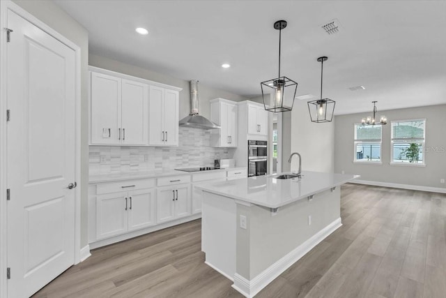 kitchen featuring light hardwood / wood-style floors, white cabinetry, decorative light fixtures, a kitchen island with sink, and wall chimney range hood