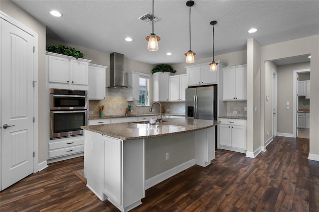 kitchen with stainless steel appliances, wall chimney range hood, an island with sink, dark stone counters, and white cabinets