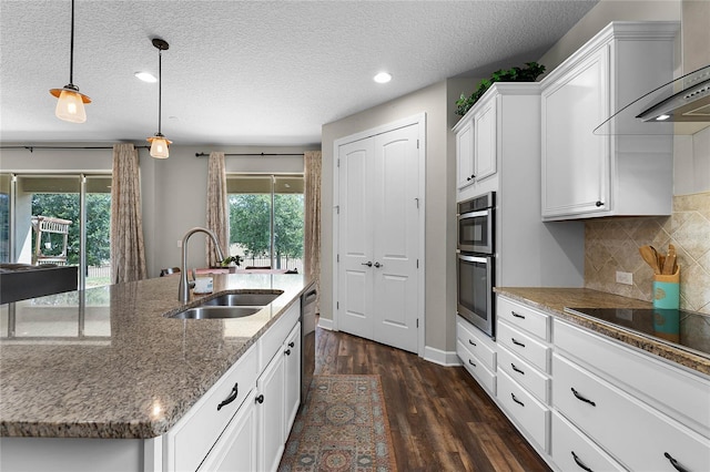 kitchen with sink, wall chimney range hood, white cabinetry, hanging light fixtures, and an island with sink