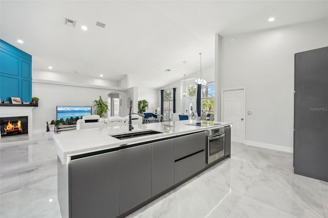 kitchen featuring black electric stovetop, stainless steel oven, a kitchen island with sink, hanging light fixtures, and sink