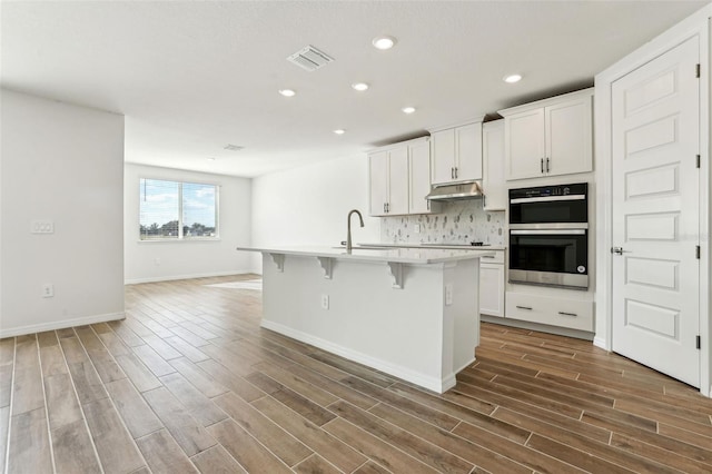 kitchen featuring tasteful backsplash, white cabinetry, an island with sink, and stainless steel double oven