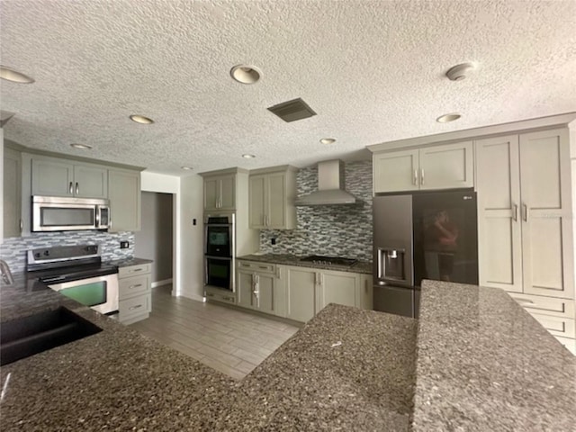 kitchen featuring appliances with stainless steel finishes, wall chimney exhaust hood, dark stone counters, sink, and backsplash