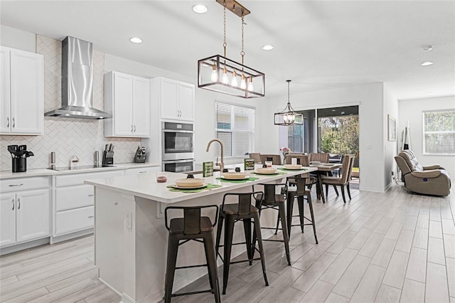 kitchen featuring a center island with sink, black electric stovetop, wall chimney range hood, double oven, and backsplash