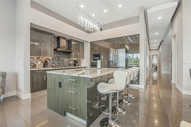 kitchen featuring a center island with sink, wall chimney range hood, light stone countertops, appliances with stainless steel finishes, and a kitchen bar