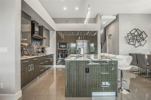 kitchen with wall chimney exhaust hood, light stone counters, a large island with sink, a kitchen bar, and appliances with stainless steel finishes