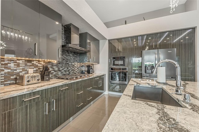 kitchen featuring decorative backsplash, light stone countertops, wall chimney exhaust hood, stainless steel appliances, and sink