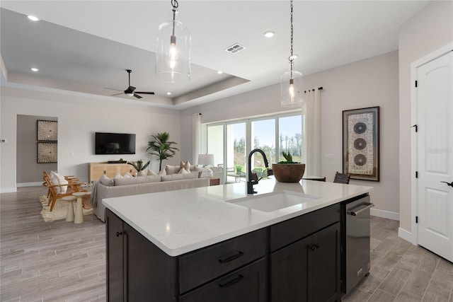 kitchen featuring a raised ceiling, a kitchen island with sink, sink, and hanging light fixtures