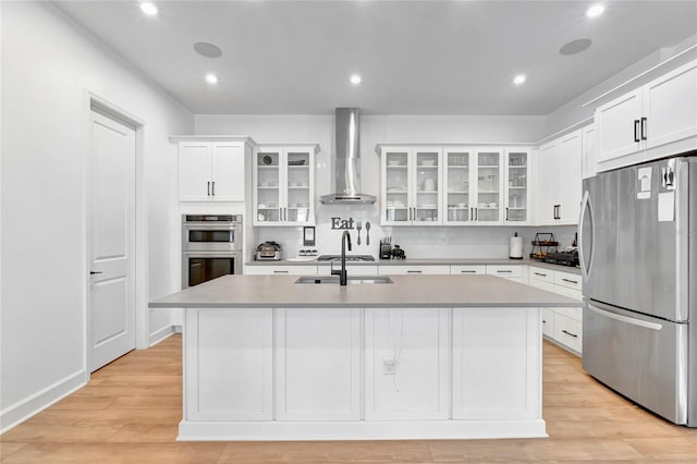 kitchen with white cabinets, wall chimney range hood, light hardwood / wood-style flooring, an island with sink, and stainless steel appliances