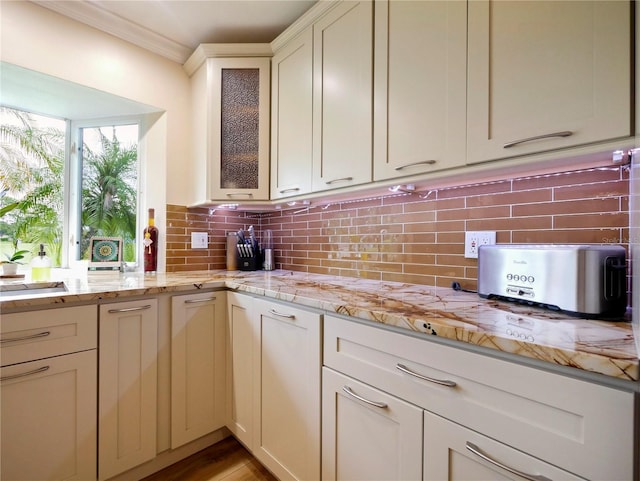 kitchen with tasteful backsplash, ornamental molding, glass insert cabinets, and light stone countertops