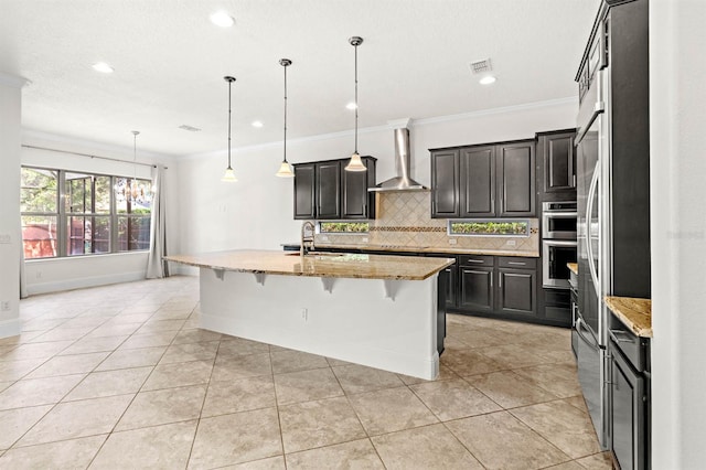 kitchen featuring a center island with sink, wall chimney range hood, light stone counters, and decorative light fixtures