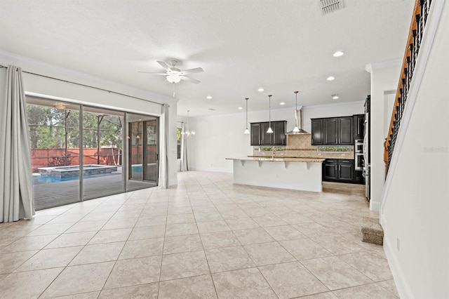 kitchen with a breakfast bar, ceiling fan with notable chandelier, hanging light fixtures, wall chimney exhaust hood, and a center island with sink