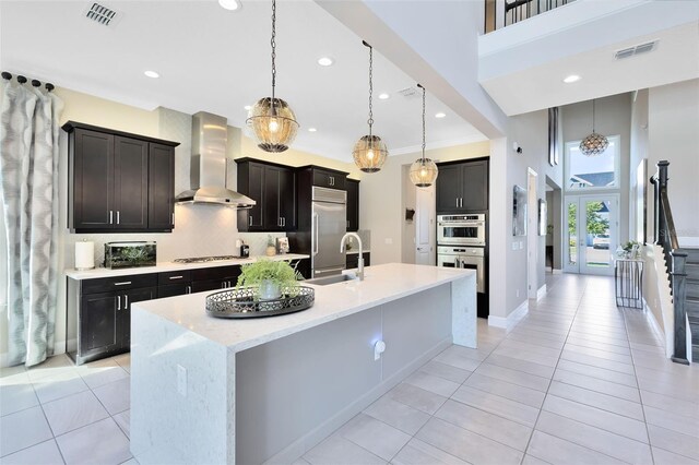 kitchen with sink, light tile patterned floors, stainless steel appliances, an island with sink, and wall chimney exhaust hood