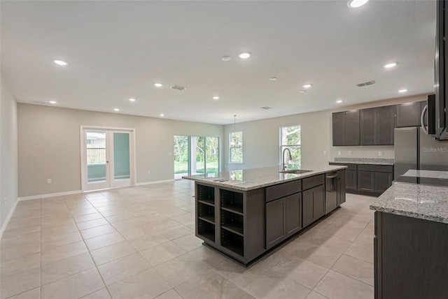 kitchen with sink, appliances with stainless steel finishes, dark brown cabinets, light stone countertops, and an island with sink