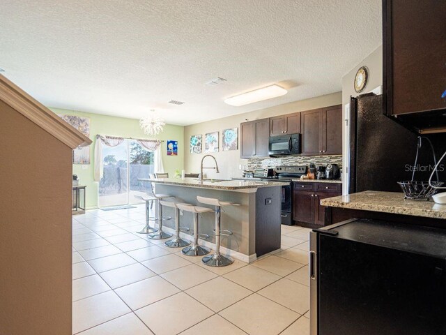 kitchen featuring an island with sink, light tile flooring, black appliances, backsplash, and a breakfast bar