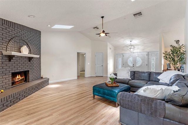 living room with lofted ceiling with skylight, a brick fireplace, ceiling fan, a textured ceiling, and light hardwood / wood-style floors