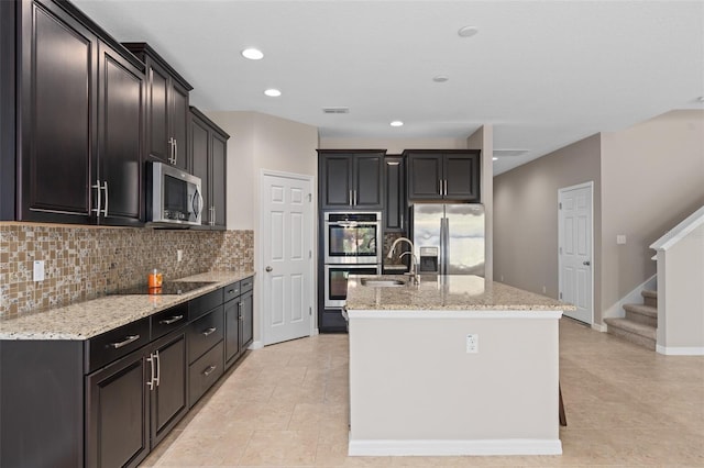 kitchen with tasteful backsplash, sink, a kitchen island with sink, appliances with stainless steel finishes, and light stone countertops