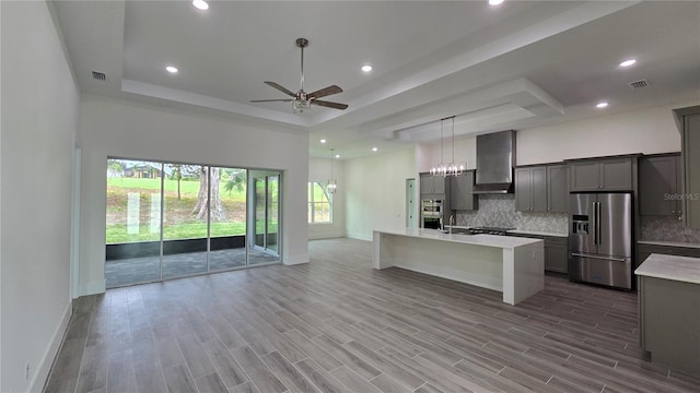 kitchen with wall chimney range hood, a raised ceiling, gray cabinets, a center island with sink, and appliances with stainless steel finishes