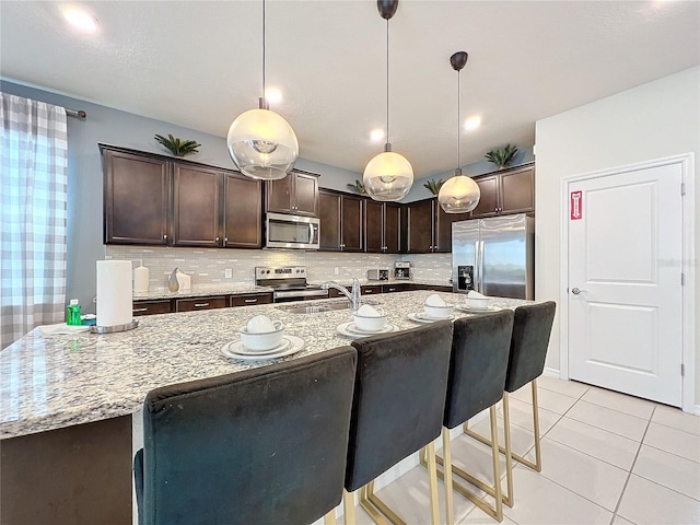 kitchen featuring hanging light fixtures, tasteful backsplash, dark brown cabinets, light stone counters, and stainless steel appliances