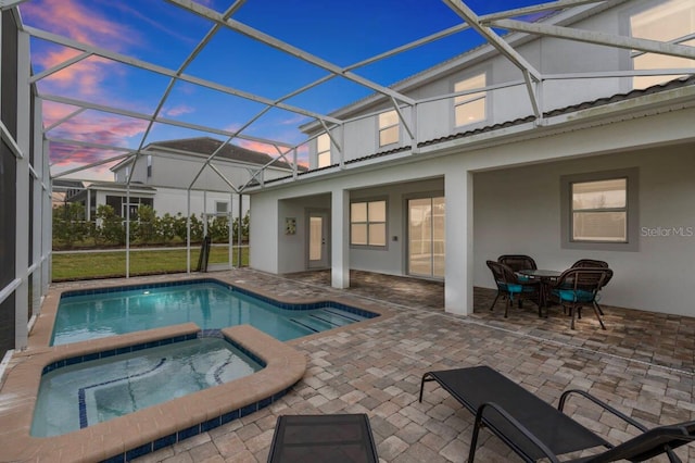 pool at dusk with an in ground hot tub, glass enclosure, and a patio area