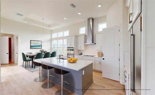 kitchen featuring wall chimney exhaust hood, a center island with sink, white cabinetry, hanging light fixtures, and a breakfast bar area