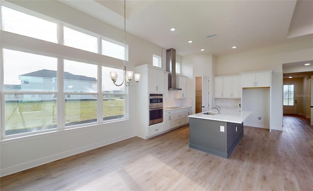 kitchen featuring pendant lighting, white cabinets, wall chimney range hood, light hardwood / wood-style flooring, and an island with sink