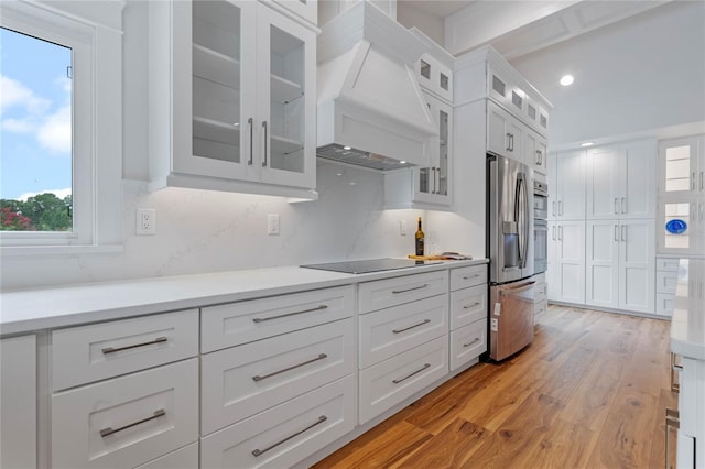 kitchen featuring stainless steel refrigerator with ice dispenser, black electric stovetop, premium range hood, white cabinets, and light hardwood / wood-style floors