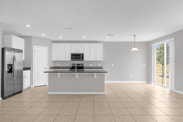 kitchen with light tile patterned floors, dark stone countertops, hanging light fixtures, a kitchen island with sink, and stainless steel appliances