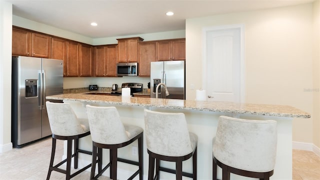 kitchen featuring stainless steel appliances, an island with sink, light stone counters, light tile patterned floors, and a kitchen breakfast bar