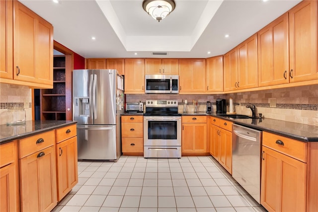 kitchen with appliances with stainless steel finishes, a raised ceiling, sink, light tile patterned floors, and dark stone countertops