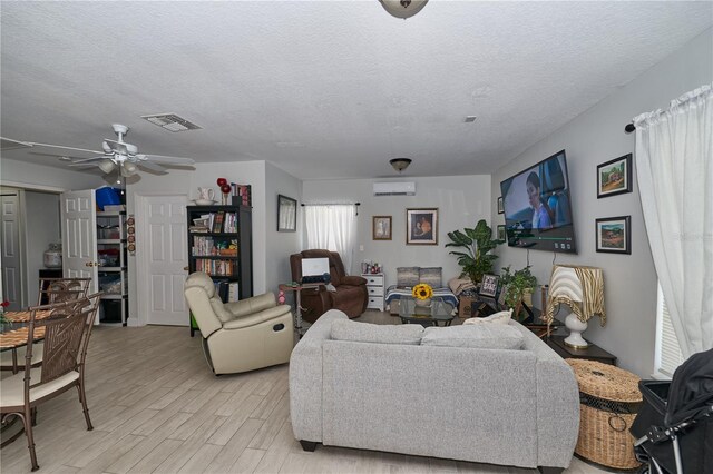 living room with a wall mounted air conditioner, a textured ceiling, ceiling fan, and light wood-type flooring