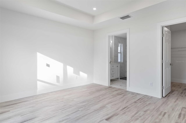 unfurnished bedroom with connected bathroom, a spacious closet, light hardwood / wood-style floors, a tray ceiling, and a closet