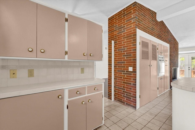 kitchen with light tile patterned floors, lofted ceiling with beams, wall oven, and brick wall