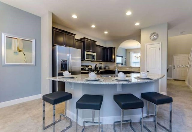 kitchen with dark brown cabinets, stainless steel appliances, an island with sink, light stone counters, and a breakfast bar area