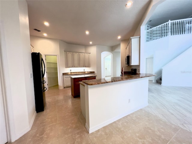 kitchen with kitchen peninsula, dark stone countertops, light tile patterned flooring, and black refrigerator