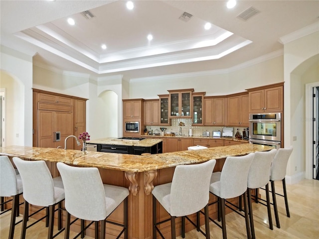 kitchen with light stone counters, a breakfast bar, a raised ceiling, a large island with sink, and built in appliances