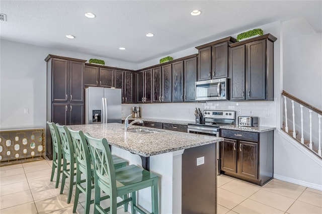 kitchen featuring a center island with sink, stainless steel appliances, dark brown cabinets, light stone countertops, and sink