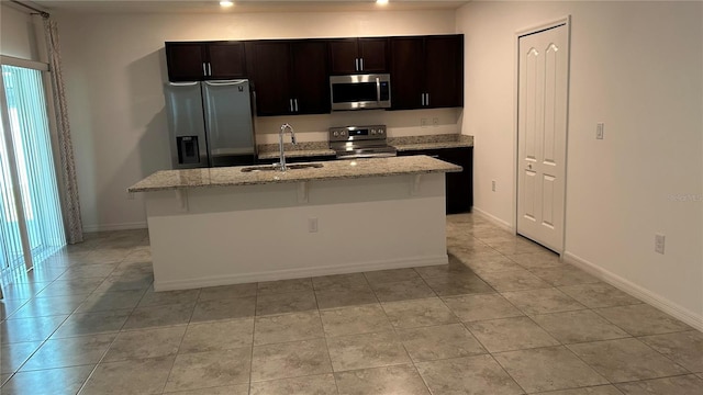 kitchen with a center island with sink, sink, light stone countertops, stainless steel appliances, and dark brown cabinetry