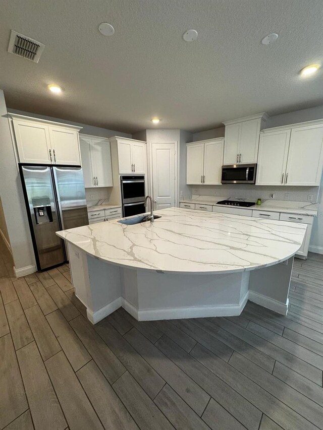 kitchen with sink, appliances with stainless steel finishes, white cabinetry, a kitchen island with sink, and light stone countertops