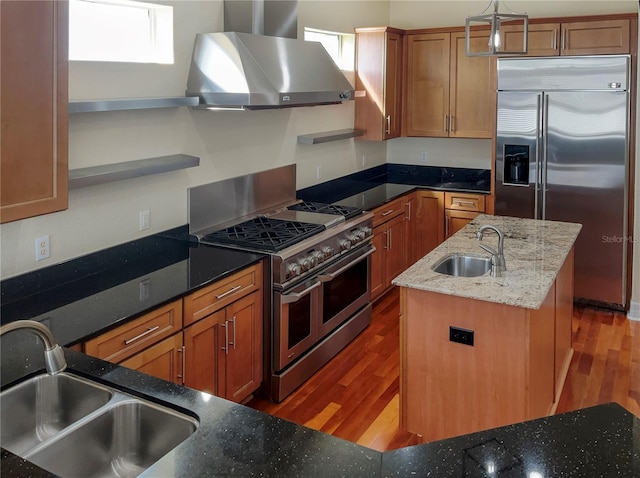 kitchen with dark stone counters, a kitchen island with sink, high end appliances, sink, and wall chimney range hood