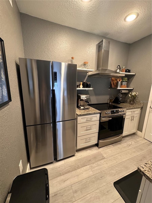 kitchen with wall chimney range hood, stone counters, white cabinetry, stainless steel appliances, and light wood-type flooring