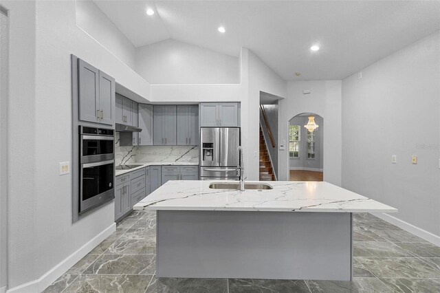 kitchen with gray cabinetry, light stone countertops, stainless steel appliances, high vaulted ceiling, and a center island with sink