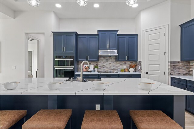 kitchen featuring decorative backsplash, blue cabinetry, light stone counters, multiple ovens, and a kitchen breakfast bar