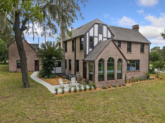 rear view of house featuring a shingled roof, a lawn, a patio, a chimney, and brick siding