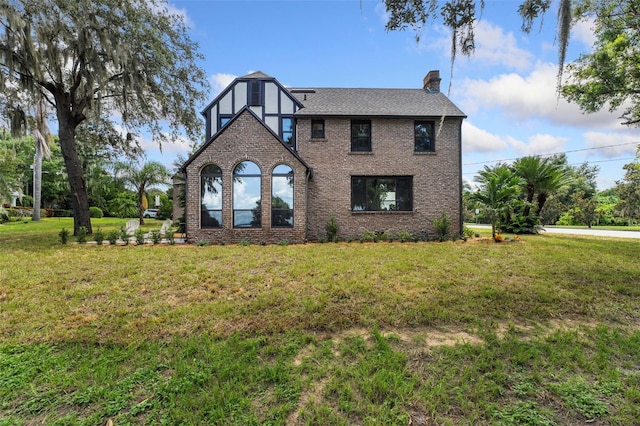 tudor-style house featuring brick siding, a chimney, a front lawn, and roof with shingles