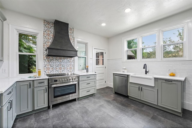 kitchen with custom exhaust hood, stainless steel appliances, gray cabinets, light countertops, and a sink