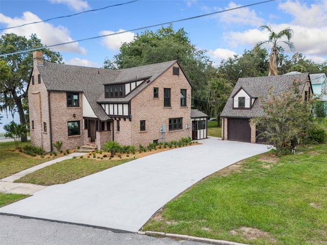 tudor house with roof with shingles, a detached garage, brick siding, an outdoor structure, and a front lawn