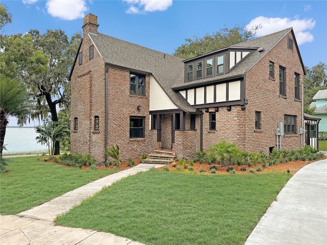 tudor-style house featuring a shingled roof, a front yard, brick siding, and a chimney