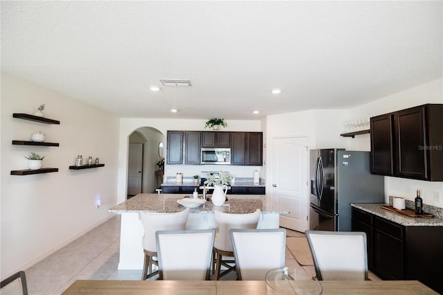 kitchen featuring a kitchen bar, appliances with stainless steel finishes, a center island with sink, dark brown cabinetry, and light stone countertops